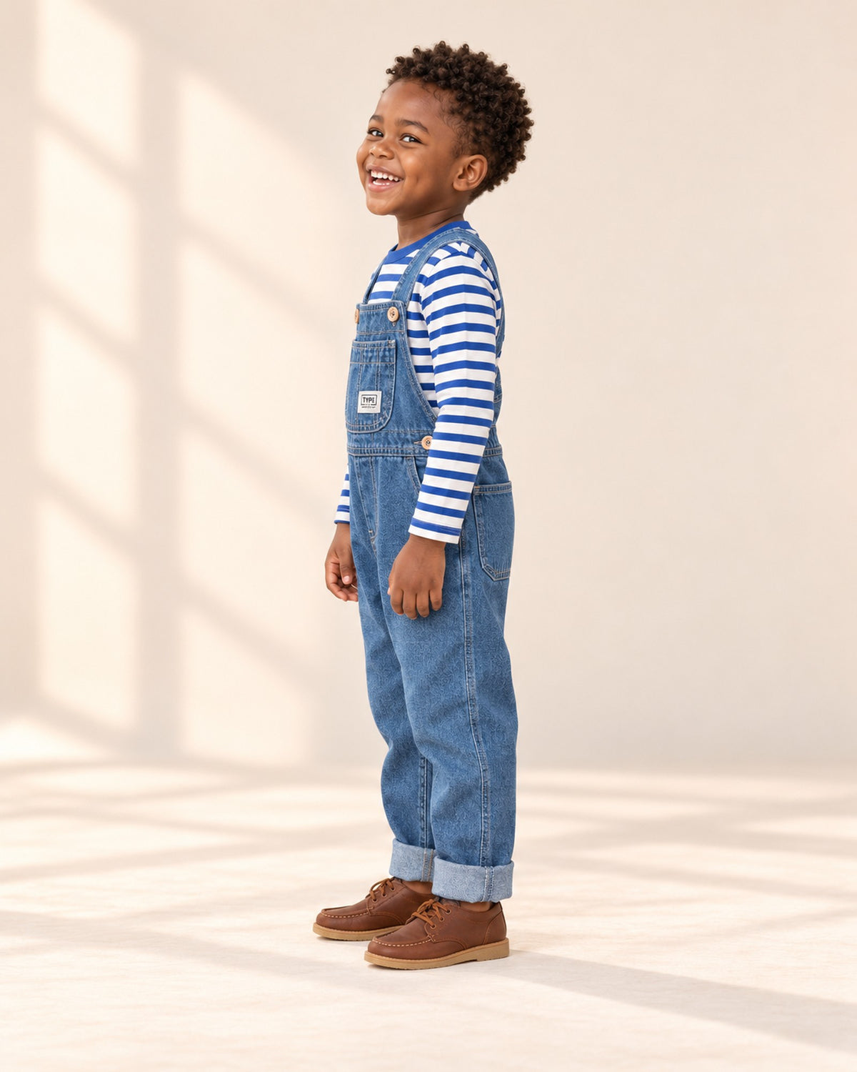 Side view of the denim overalls showing the strap and cuffed leg over the striped tee – Mid Wash