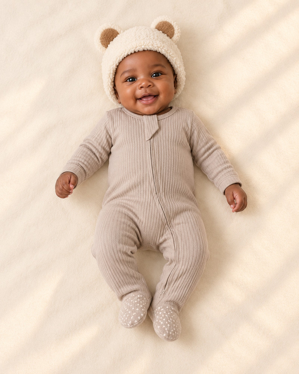 Baby in the cream bear ear beanie and a taupe ribbed zip footie, full length – Cream