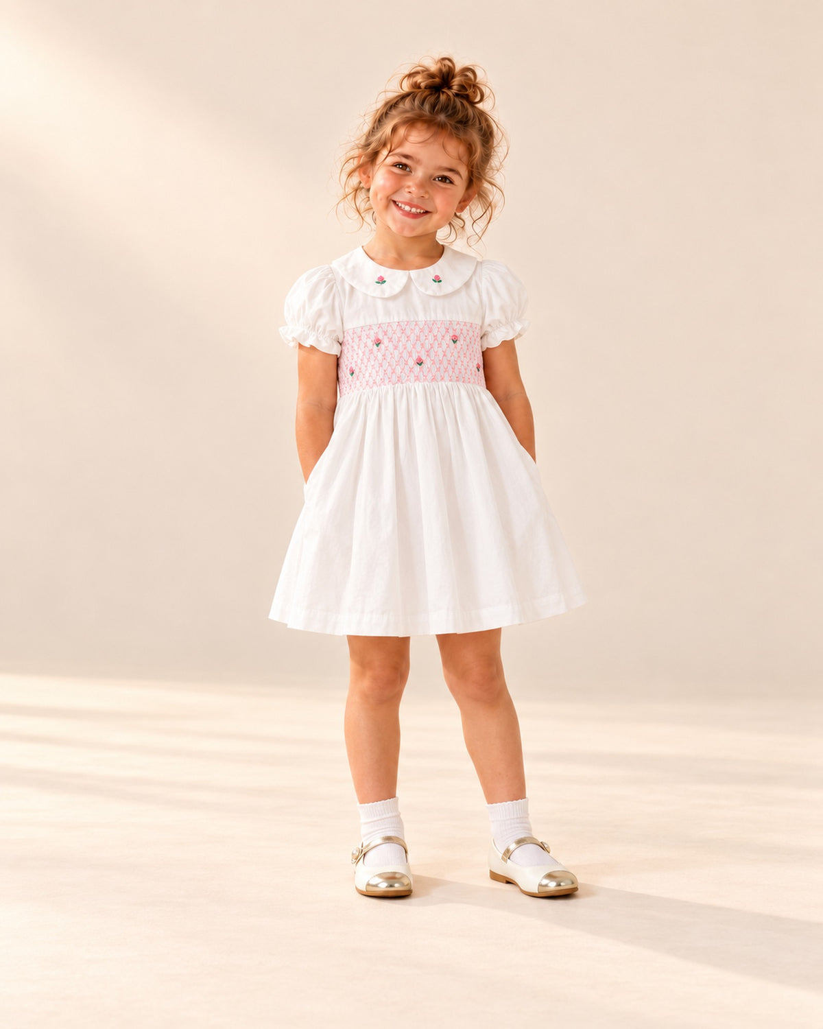 Front view of a girl in a white rosebud smock dress, white ankle socks and pearl strap Mary Janes, full length – White