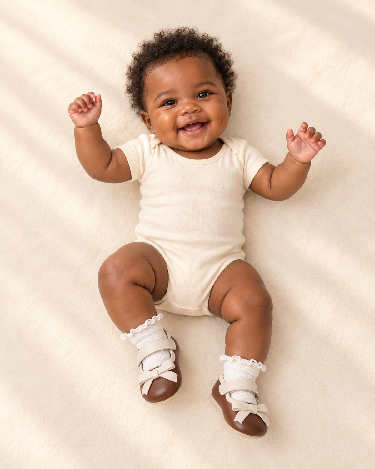 Front view of a baby in a plain cream romper, white frill socks and brown bow pre-walkers, full length – White