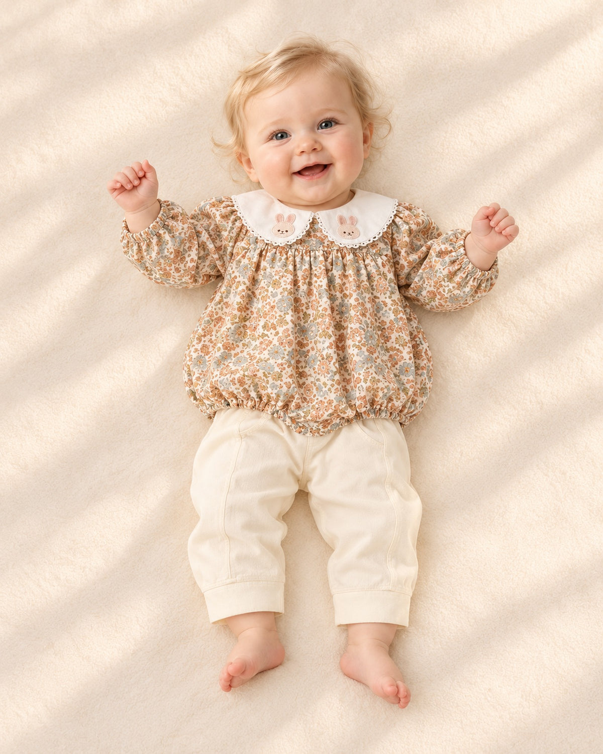 Front view of a baby in the cream pants under a floral bubble romper with a bunny collar, full length – Cream