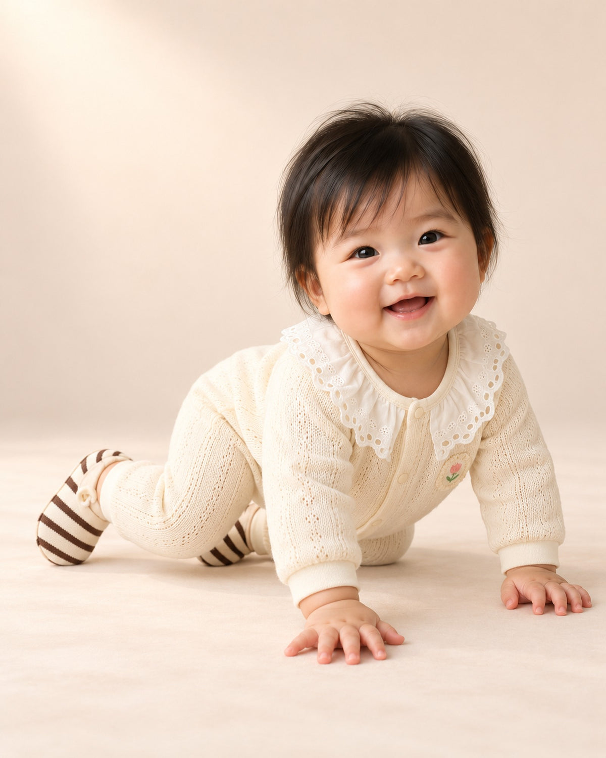 Baby sitting on a blanket in a cream knit romper and striped sock shoes – Brown Stripe